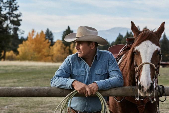 kevin costner u Yellowstoneu pokraj konja naslonjenog na ogradu s užetom u rukama nosi izblijedjelu plavu traper košulju i bež kaubojski šešir