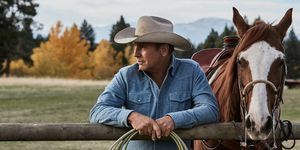 kevin costner u Yellowstoneu pokraj konja naslonjenog na ogradu s užetom u rukama nosi izblijedjelu plavu traper košulju i bež kaubojski šešir
