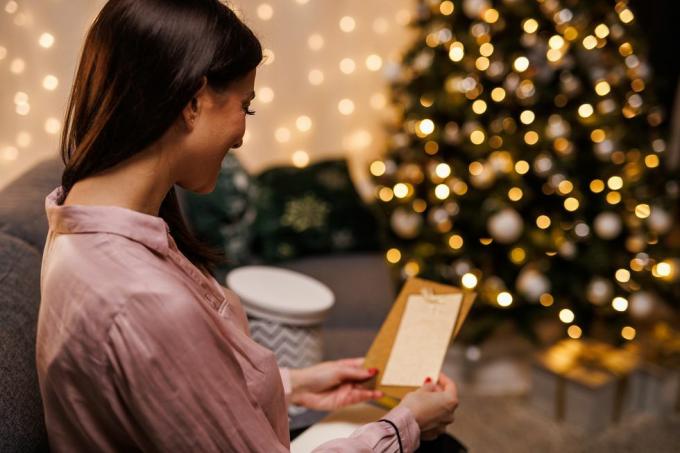sretna mlada žena sjedi na sofi, pokraj svjetlucavog božićnog drvca, otvara i čita božićnu čestitku koju je dobila s poklonom