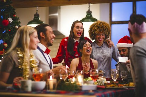 grupa prijatelja koji se smiju dok sjede za stolom i jedu i piju za Božić jedan čovjek čita vic s božićnog krekera nose se šeširi i božićni džemperi